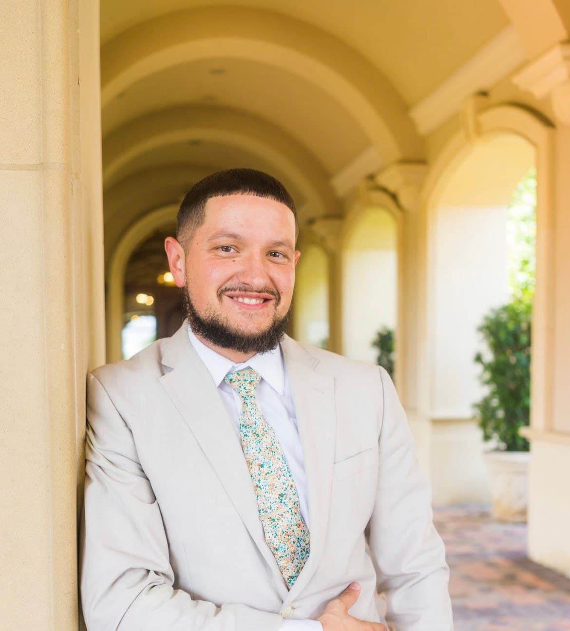 David Settle in a light suit and patterned tie, leaning against a column in an arched walkway, smiling